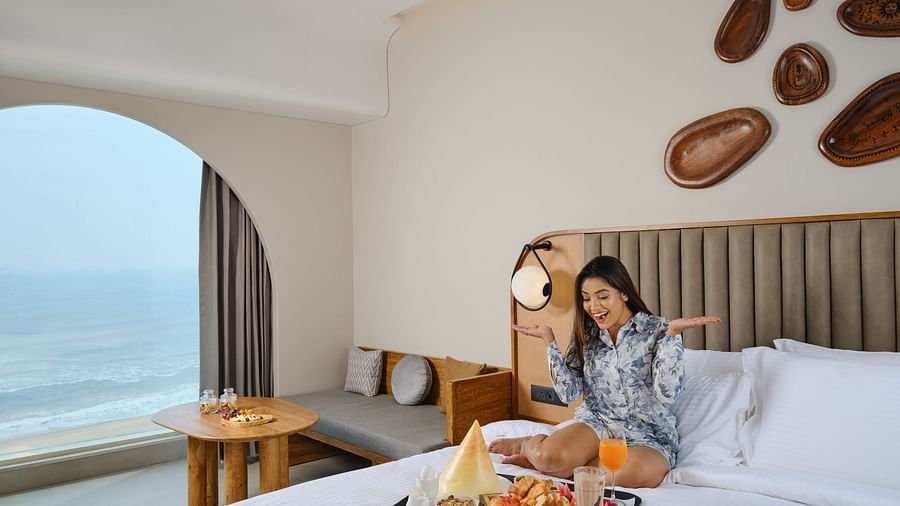 A woman excitedly enjoying breakfast in bed at Swosti Premium Beach Resorts, Puri, in the Sea View Room, which boasts modern decor and a stunning ocean view through an arched window