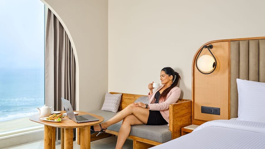 A woman relaxing in the Sea View Room at Swosti Premium Beach Resorts, Puri, featuring modern decor, a cozy seating area, and a ocean view through an arched window