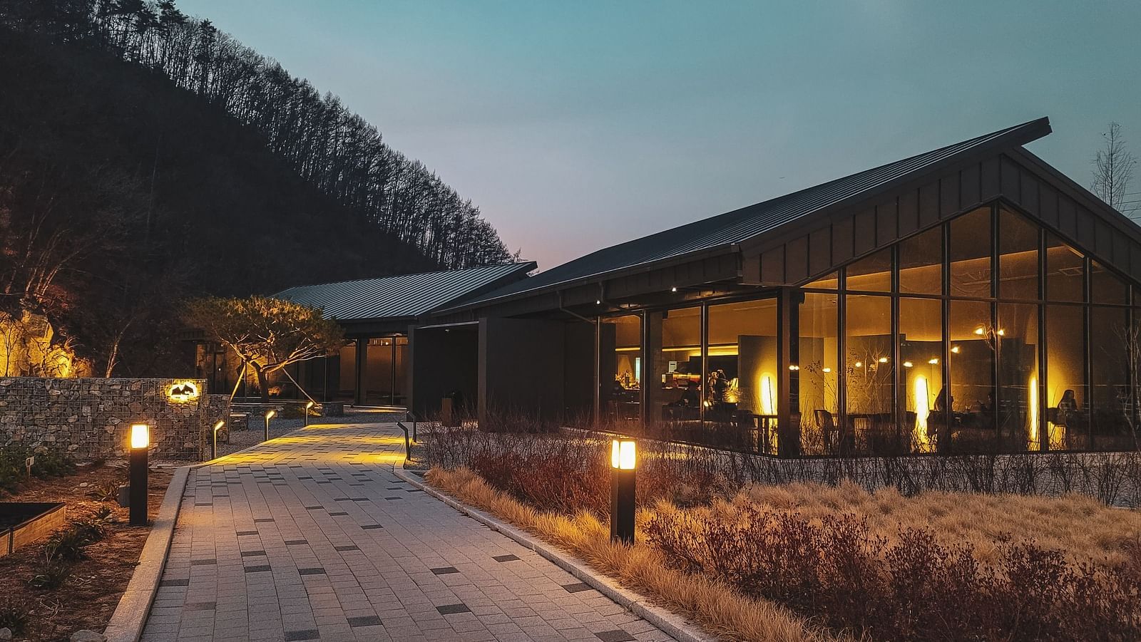 A modern building with glass walls illuminated from within at twilight with a pathway lined by lit lamps - The Tanisi Hotels & Resorts, Goa