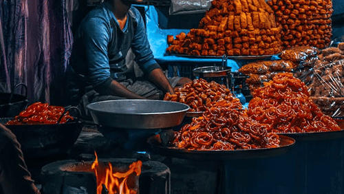 A person sits behind a stall with piles of food items and a stove with a flame visible below.