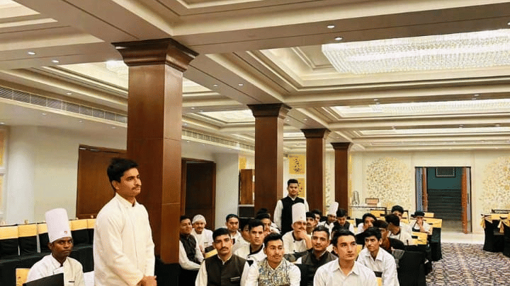 People gather inside a hall at Clarks Group of Hotels. Tables and chairs are arranged across the room for an event.