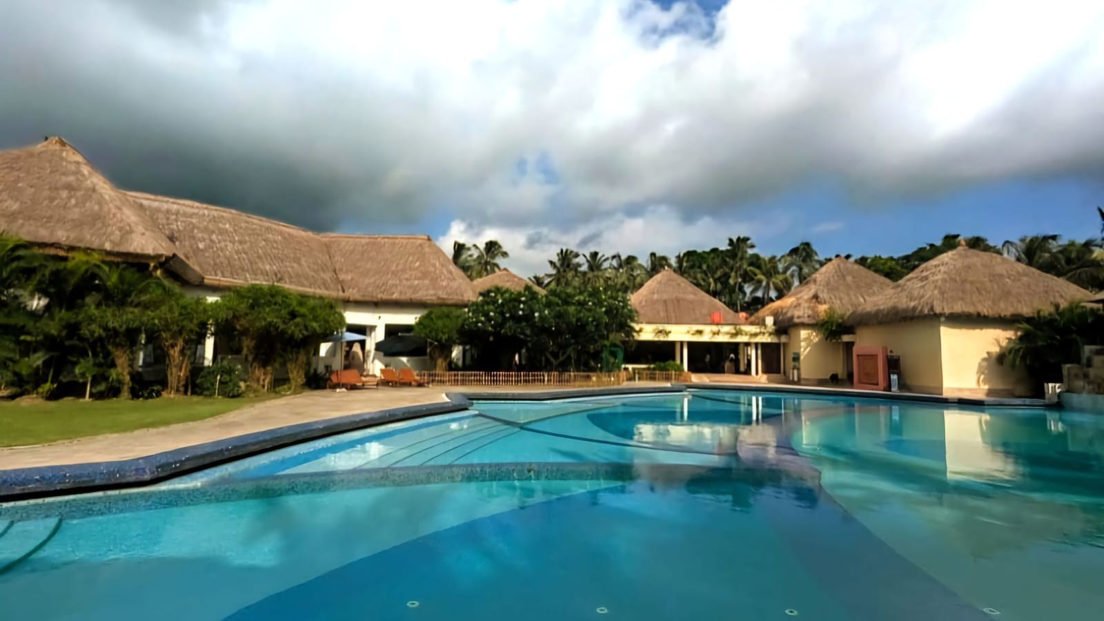 A wide view shot of Vedic Village Spa Resort featuring a swimming pool lined by buildings with thatched roofs under a partly cloudy blue sky.