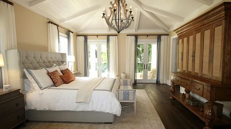 A spacious bedroom with a white bed, vaulted ceiling, chandelier, and a large wooden wardrobe.