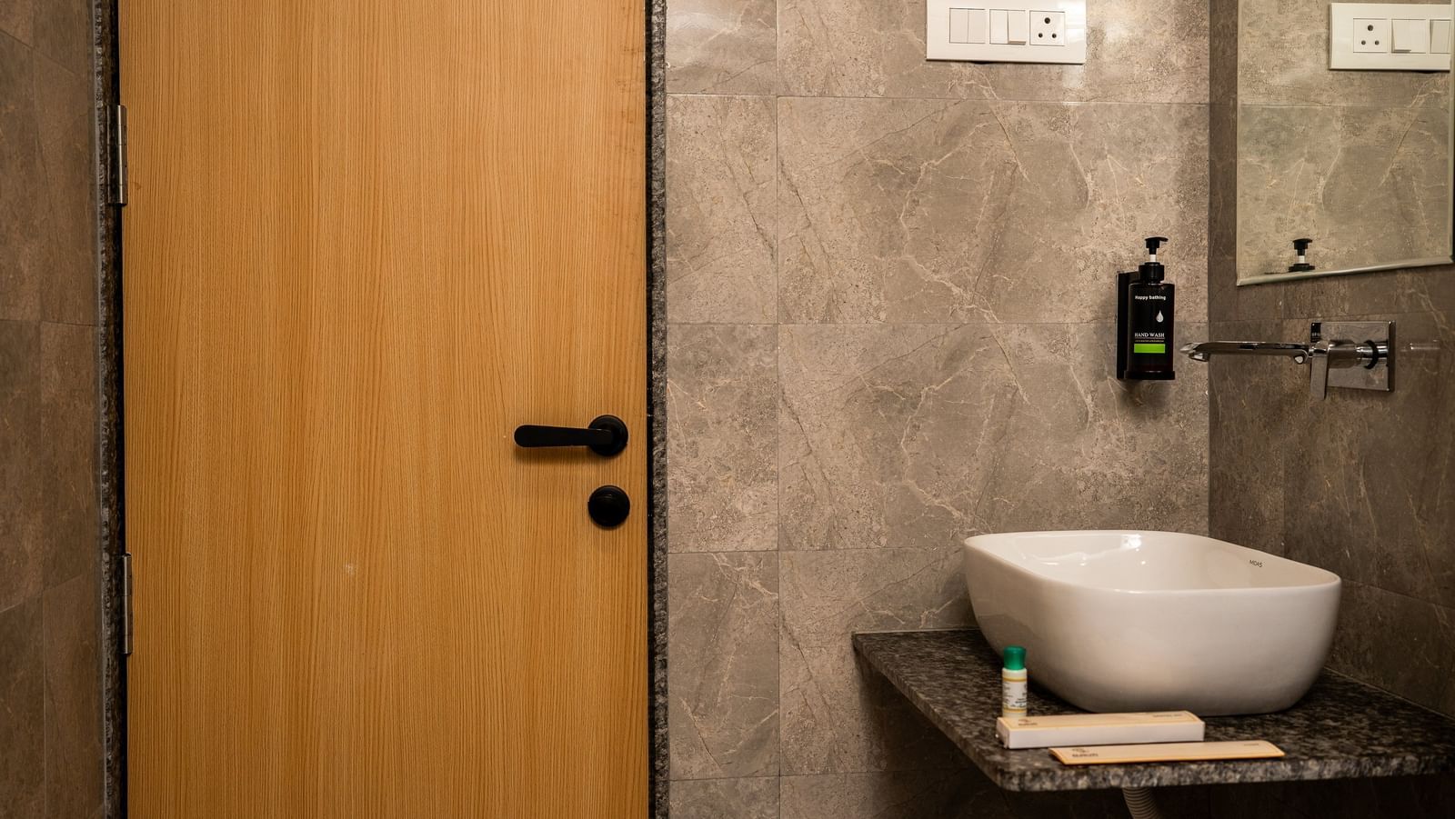 A contemporary bathroom at Daksh The Grand Gowardhan, Dwarka, with a sleek basin and wooden door besides it.