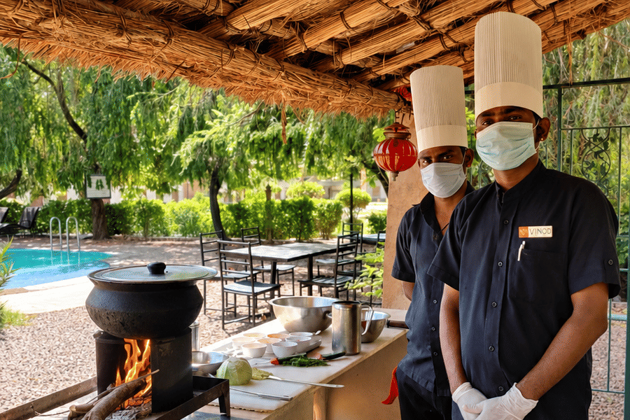 Two chefs in hats and masks standing at an outdoor live cooking station with a pot over a wood-fire stove under a thatched roof near a pool.
