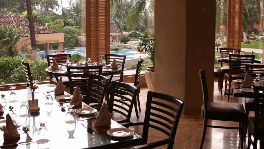 A restaurant dining area with wooden chairs and tables overlooking  a pool area framed by palm trees.