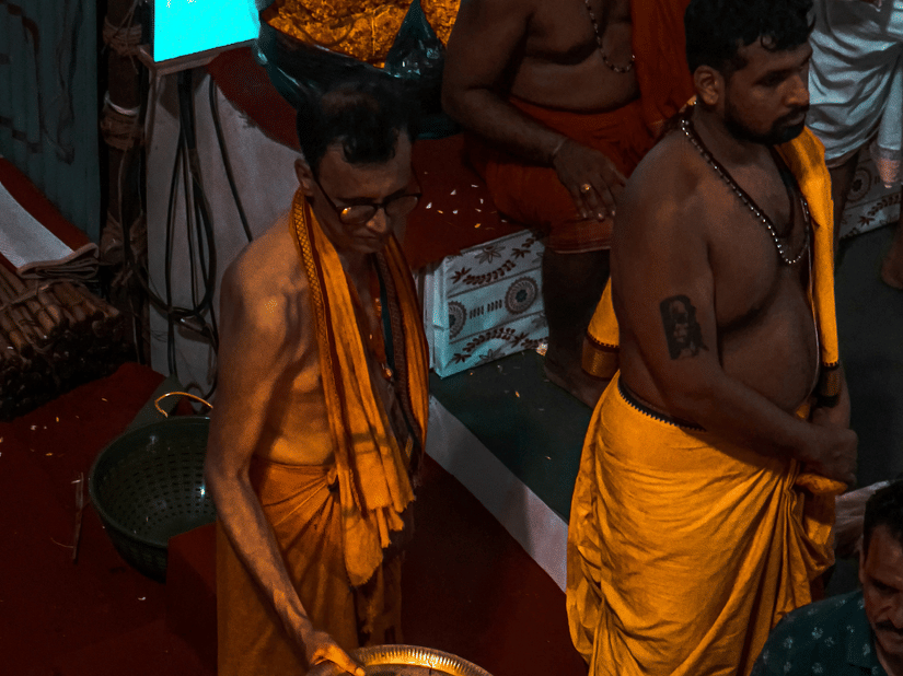 A view from above of two temple priests performing rituals. One chanting prayers and another holding a brass plate with a lit fire.