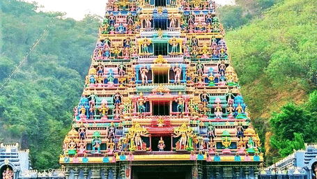 A vibrant facade of the Kanaka Durga Temple in Vijayawada featuring a multi-tiered temple gopuram against a green and hilly backdrop.