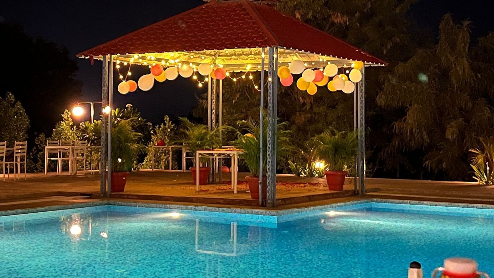 A nighttime shot of a gazebo with a red roof, decorated with lights and balloons, next to a brightly lit swimming pool - Bamboo Saa Mulberry Resort, Pushkar
