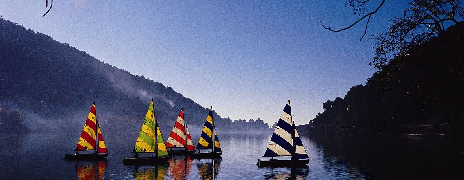 An image with five sailboats pictured on a lake , with their reflections visible on the water, with the magnificent backdrop of the mountains converging.