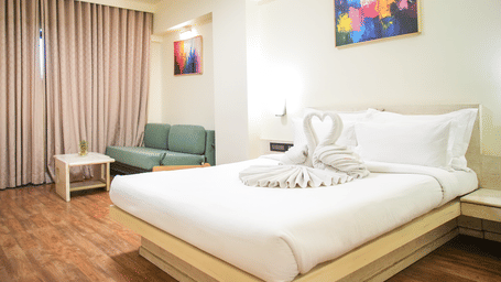 A room with a king size bed, a sofa and a chair, wall arts and light brown curtains at Infiniti Hotel, Indore.