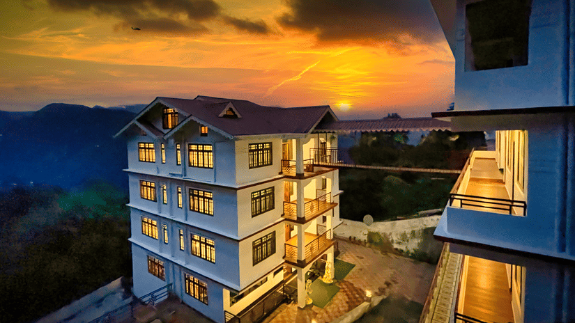 Voyage The Royal Retreat, a hillside hotel illuminated with warm yellow lights at night, set against a dramatic deep blue cloudy sky with a paved driveway and mountain silhouettes surrounding the property.