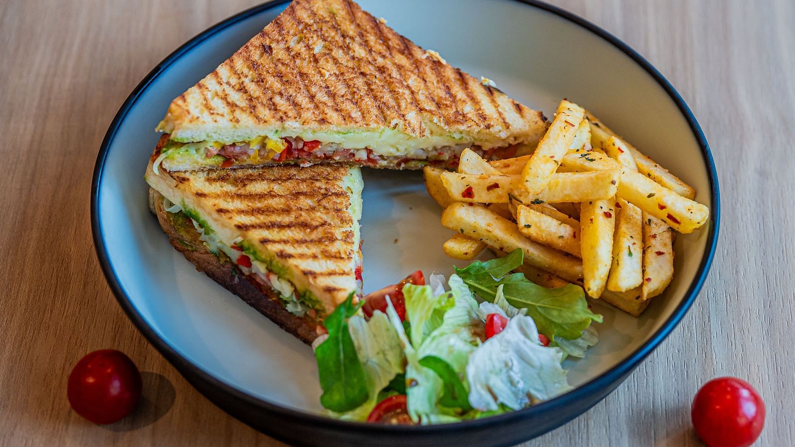 A plate with two triangular slices of sandwiches, accompanied by vegetables on the side, along with a pile of fries and mini tomatoes placed beside the plate at Indie Stays Mumbai International Airport.