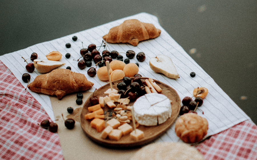 Picnic spread of bread, fruit and cheese on a red checked cloth with a straw hat beside still water