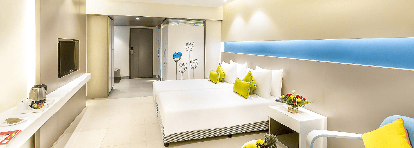 A spacious room features a large bed with a headboard stretching across the wall. Cushion chairs sit beside a wooden bedside table, and a wall-mounted television is positioned opposite the bed