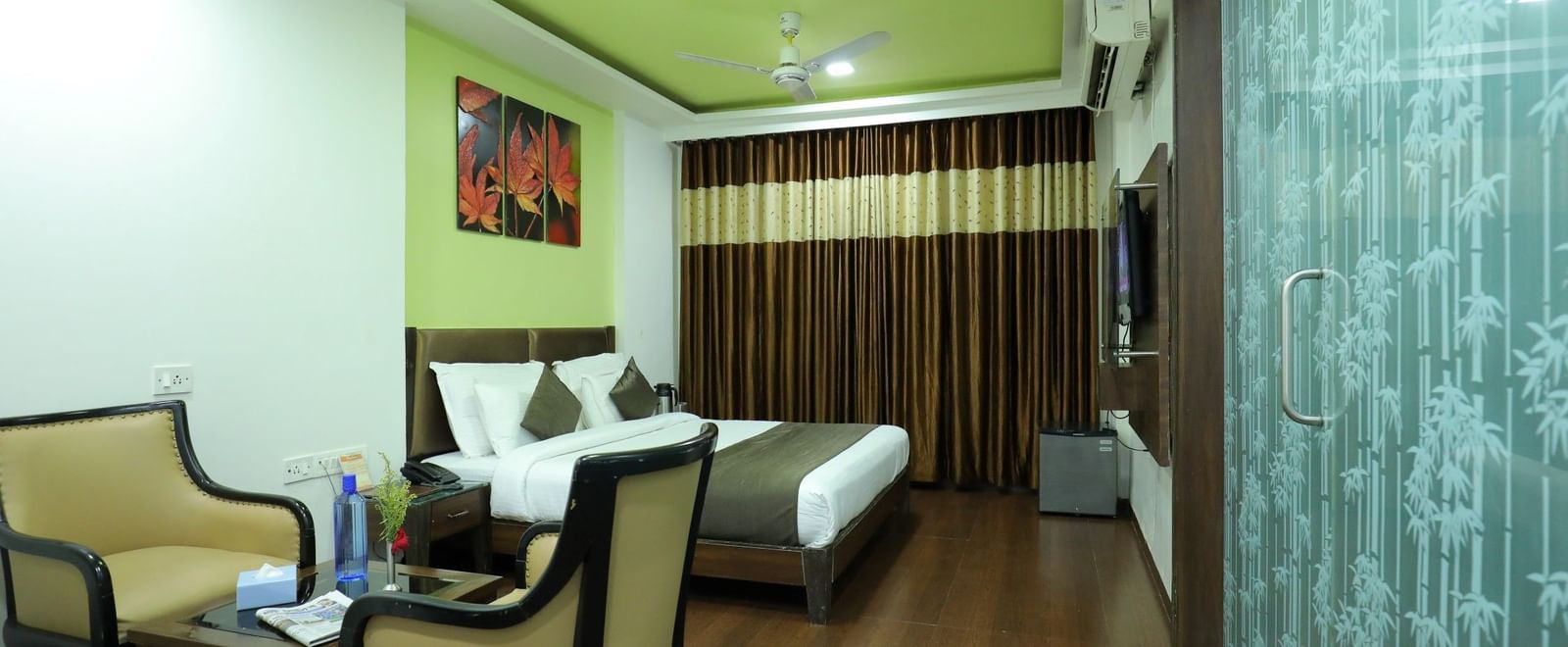 A bedroom at Krushnai Resort with a bed, 2 chairs, a glass parting on one side, and a vibrant green ceiling.