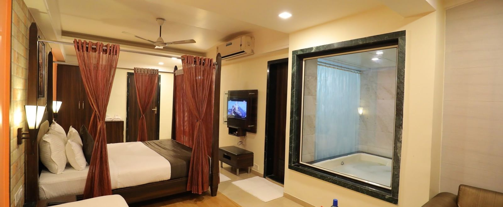 A suite at Krushnai Resort featuring a four-poster bed, a separate seating area, and a glass panelled wall with a view of a bathtub.