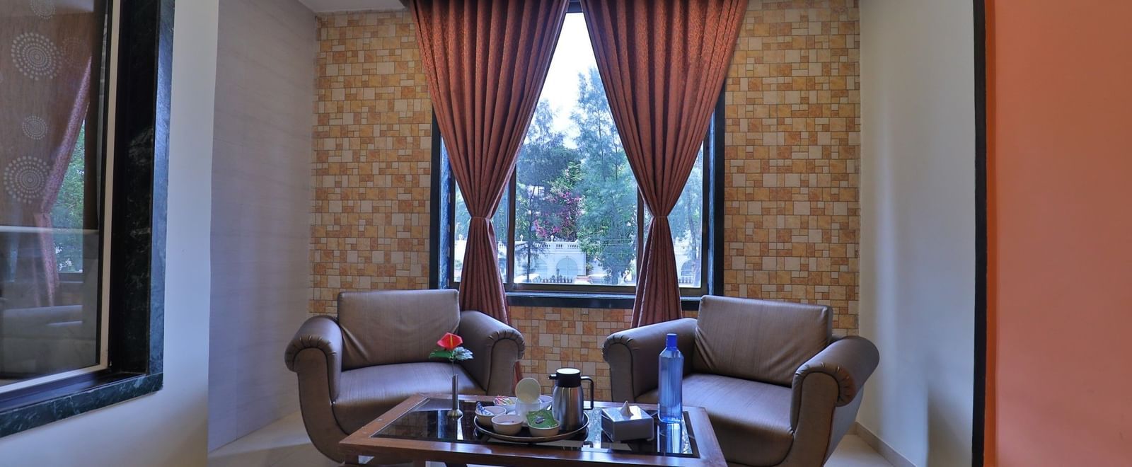 A seating area in a suite at Krushnai Resort with 2 lounge chairs, coffee table, and red drapes, beside a window.
