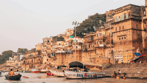 The view of Varanasi ghats with historic riverside buildings, boats, and people along the Ganges River.