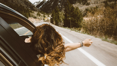 A person extending their hand out of a car window while the vehicle drives along a road with mountains in the background.