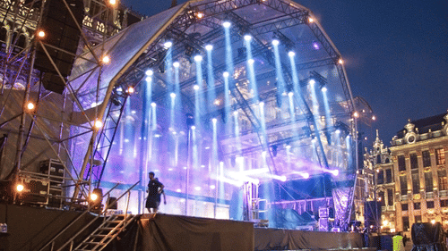Outdoor stage in a city square illuminated with vibrant purple and blue lights.