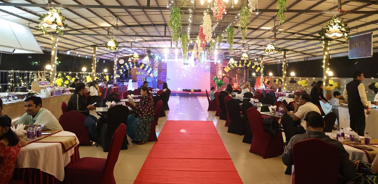 An outdoor event space is decorated with lights and a red carpet leading through seated guests - Lords Inn Vadodara