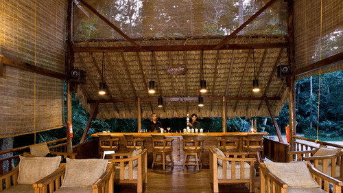 A spacious open-air wooden seating area with sofas and tables, featuring a high ceiling and large openings that overlook trees and greenery near Neil Cove Beach, Havelock, at Barefoot at Havelock.