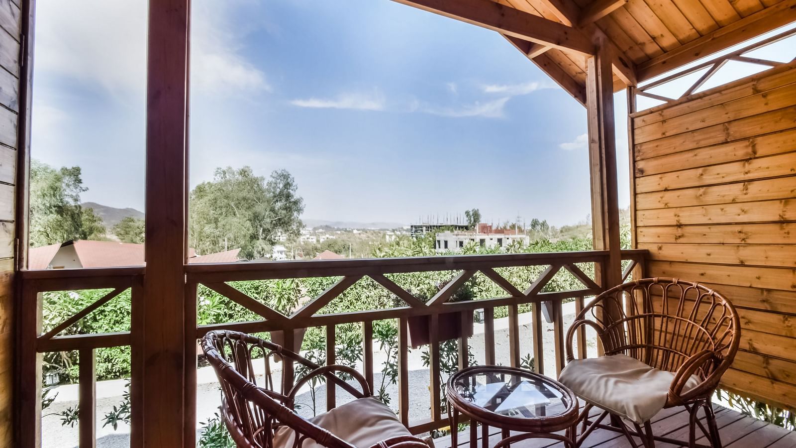 A wooden balcony with two wicker chairs and a small table overlooks a scenic view of trees .- Bamboo Saa Resort & Spa, Udaipur