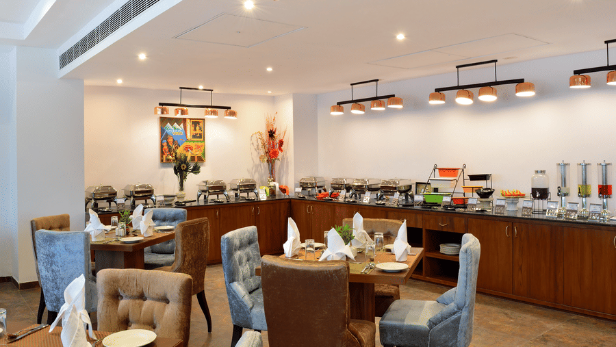 Bamboo Saa Resort & Spa—a luxury resort in Udaipur featuring a well-lit dining area with neatly arranged tables, cushioned chairs, and a wooden buffet counter with food presented in stainless steel chafing dishes.