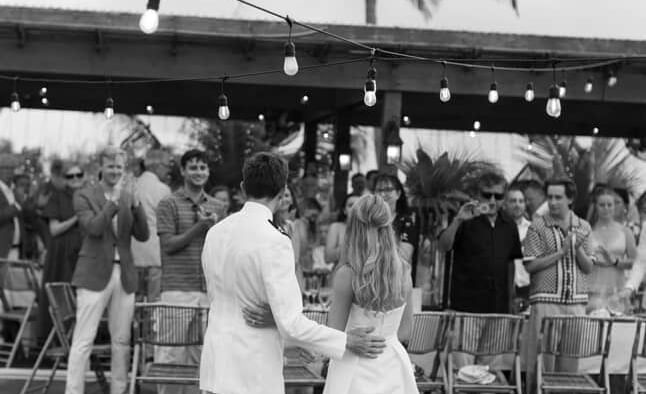 Couple holds hands facing each other on green grass while guests stand nearby, palm tree and shaded area in background at Abaco Inn.