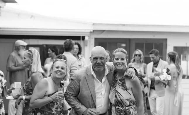 People stand on a covered terrace, some raising hands with drinks, with wedding attire, Abaco Inn.