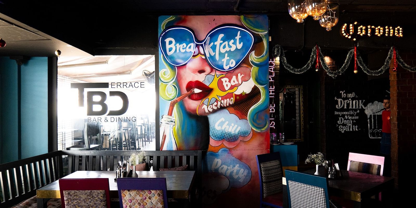 A colourful indoor dining area with a large pop art mural of a woman wearing sunglasses and drinking through a straw. The mural includes the words Breakfast to Bar. The restaurant is called TBD Bar & Dining. 