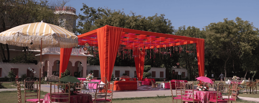 A vibrant outdoor event setup on a lawn featuring a bright orange canopy and pink table arrangements under a clear sky - Umaid Palace
