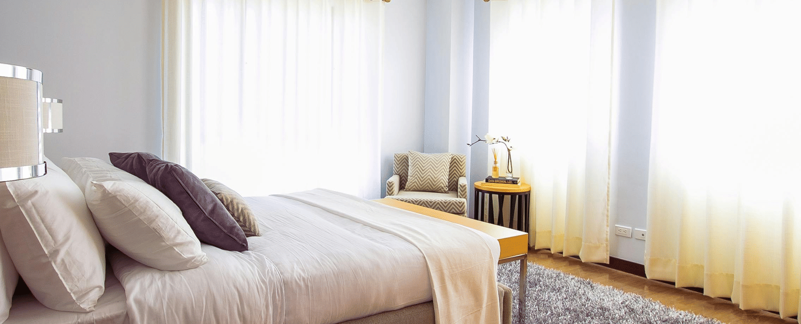 A neatly arranged bedroom with a large bed, pillows, side tables, armchair, and soft natural light filtering through curtains.