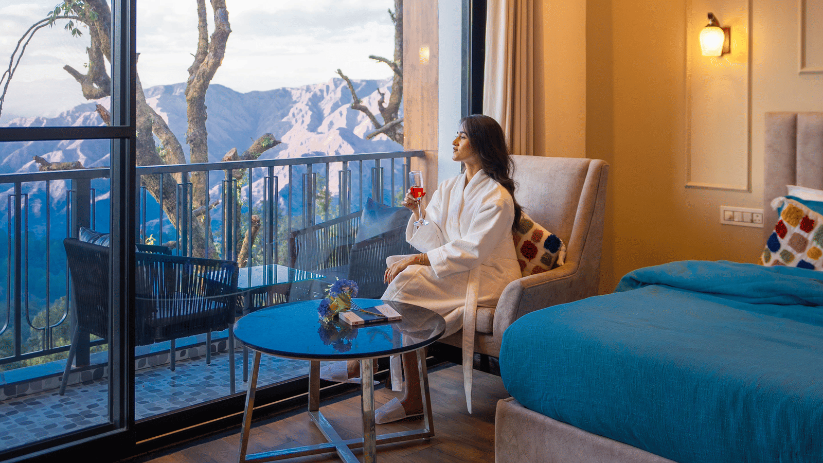 A woman dressed in a bathrobe looking outside of the Executive Room With Balcony & Valley View as she is sitting on an armchair at Perfectstayz Premium Westend Mall Road, Mussoorie.