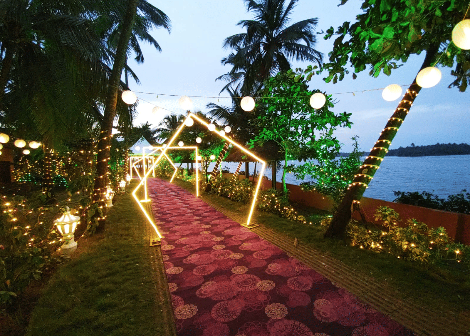 Pathway lined with lights and flowers leading towards the water with palm trees at Paradise Lagoon Resort, Udupi.