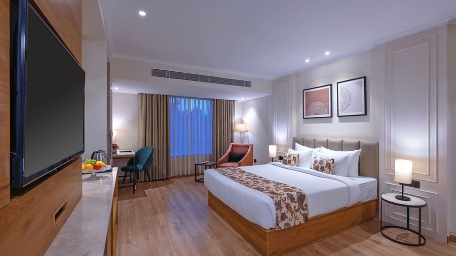 A room with a neat bed, a TV, side table lamp, wall paintings and sofa chairs - Royale Sarovar Portico, Agra