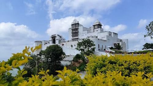 Sajjangarh Monsoon Palace, rises above manicured gardens and vibrant yellow foliage, set against a partly clouded blue sky.
