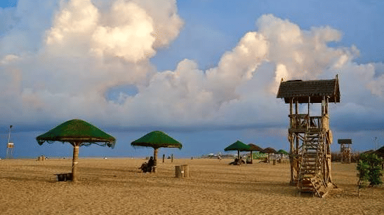 Kovalam Beach features sand structures, shelter umbrellas, and a wooden tower. Clouds fill the sky above the sea line.