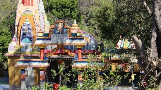 A vibrant, multicoloured temple nestled amidst dense greenery, featuring traditional Hindu motifs and deities, with a peaceful natural setting all around.