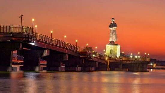 A majestic riverside statue on a lit-up bridge reflects in peaceful waters during an orange-pink evening sky in Ranchi.