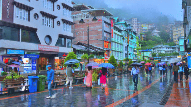 A vibrant hill station street on a rainy day with colourful buildings, pedestrians with umbrellas, and misty hills in the background at MG road.