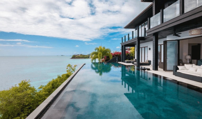 A modern luxury villa with an infinity pool overlooks a calm turquoise ocean under a partly cloudy sky.