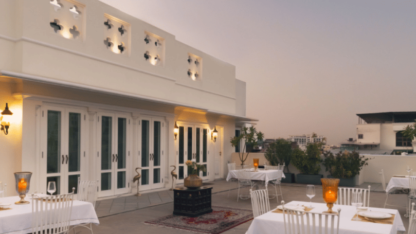 A rooftop terrace with white buildings, French doors, and outdoor dining tables set with white tablecloths, under a dusky sky, with decorative lanterns lit | Dileep  Kothi, Jaipur