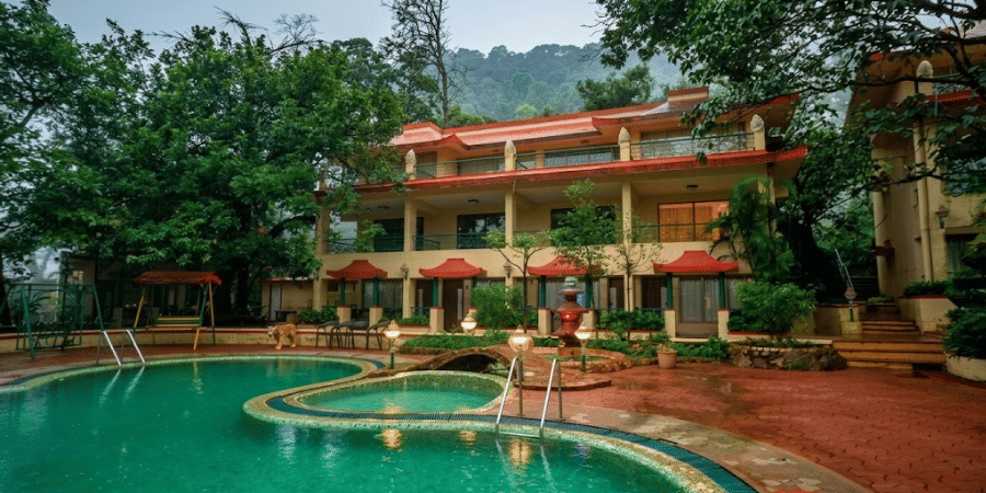 The swimming pool of the hotel with the hotel building behind it, surrounded by lush green trees.