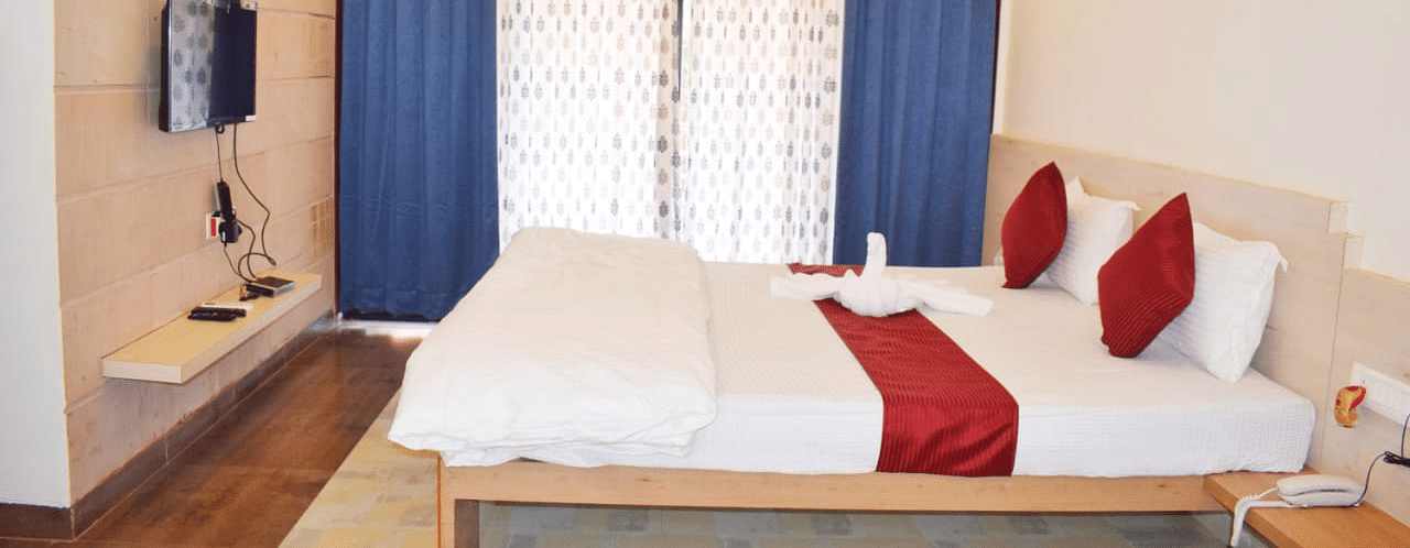 A side view of a spacious room with a large bed. Right opposite to it, there is a wall-mounted TV, and a large window with curtains fully covering it at SeaFort Beach Resort