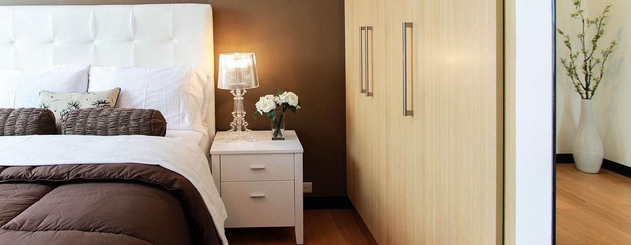 Bedroom with a bed, pillows, blanket, bedside table with lamp and flowers, and a wardrobe with sliding doors.