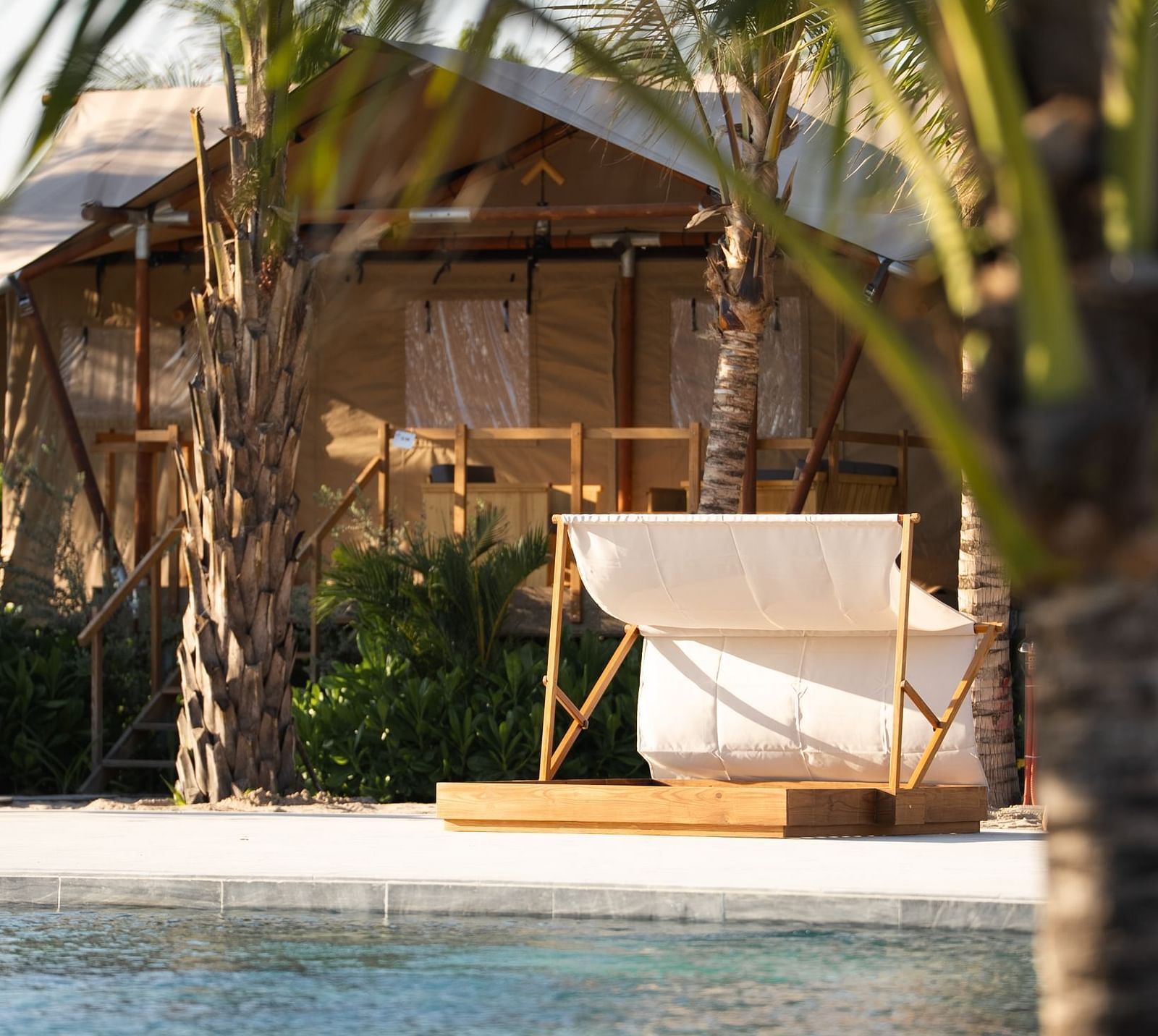A serene view of the Grateful Tent at WoCAL Retreat Ke Ga, partially framed by palm trees, with a hammock-style lounger placed beside a calm swimming pool. The tent structure in the background features a raised wooden deck and canvas covering, blending nat