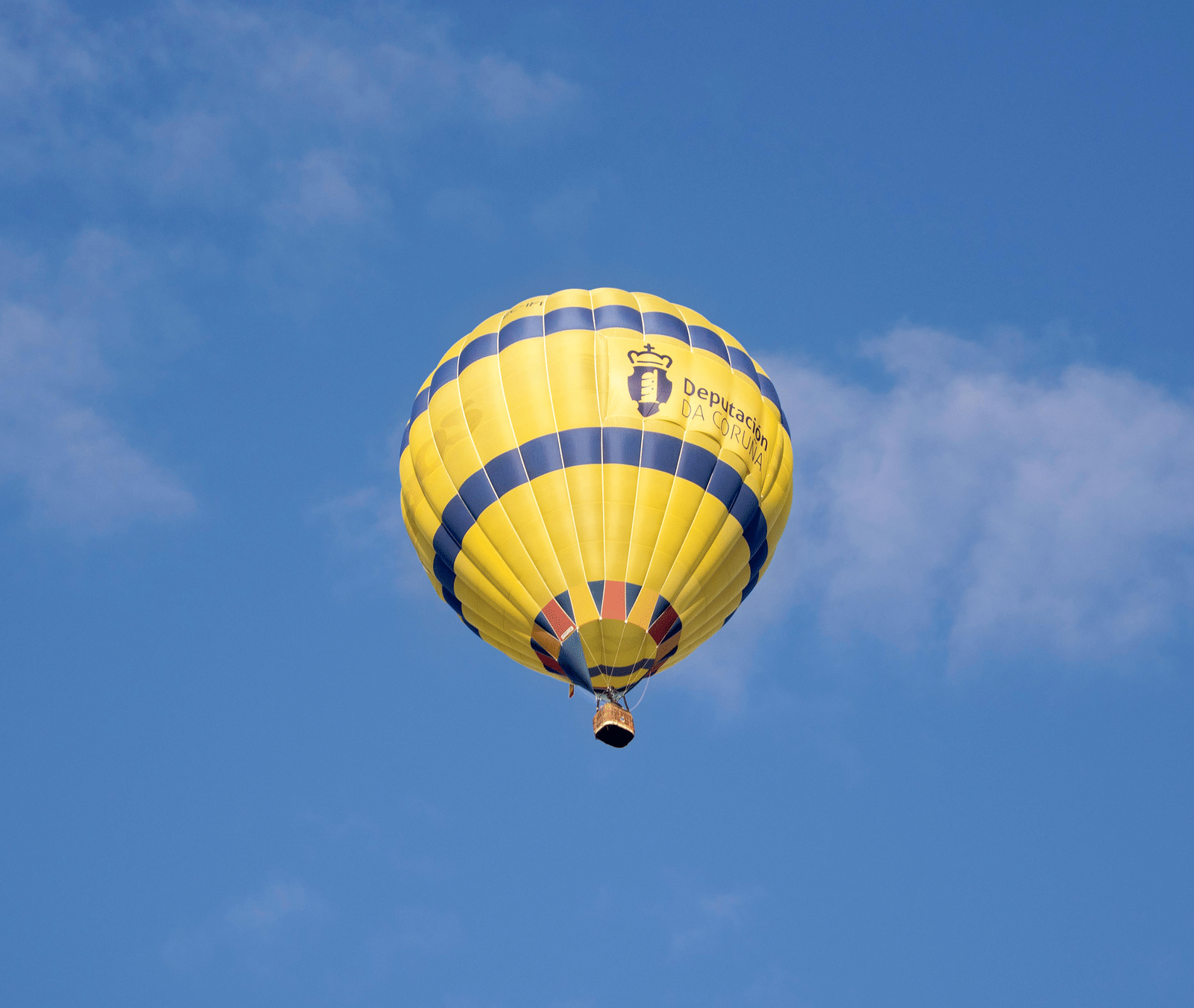 A vibrant yellow hot air balloon with 2 blue stripes flies against a bright blue sky with wispy clouds.