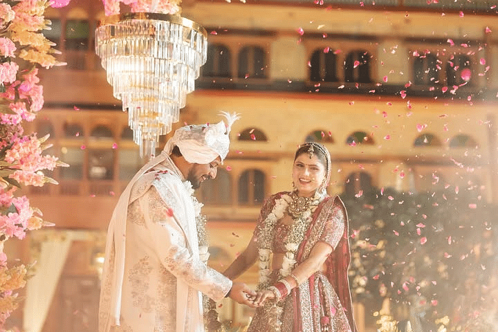A bride and groom hold hands under a floral arch as rose petals fall, with a grand palace illuminated in the background. - Umaid Palace - A Lakeside Heritage Palace Resort
